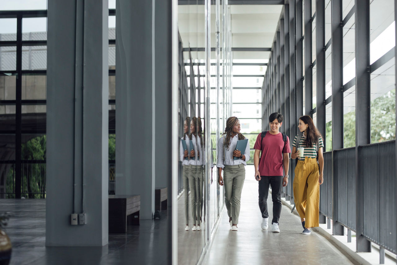 Multiethnic group of university students walking together in a hallway and looking happy. One woman is holding a folder, the other is holding a cup.